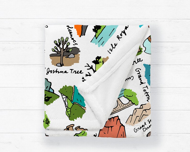 Puede incluir: Manta blanca con un dise&ntilde;o de mapa ilustrado colorido con puntos de referencia como Joshua Tree y Grand Teton. La manta tiene una textura suave y afelpada y est&aacute; doblada.