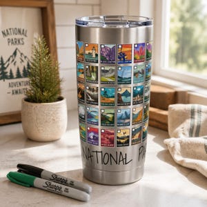 May include: A stainless steel tumbler featuring a collage of National Park images. The tumbler has a black lid and the words "NATIONAL PARKS" and "NATIONAL PARK" are printed on the side. Two Sharpie markers, black and green, are in the foreground.
