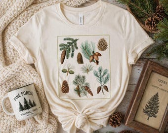 May include: Cream-colored t-shirt featuring a botanical illustration of various pine cones and branches within a green-lined square. A white mug with the text "PINE FOREST NATIONAL PARK" is visible. A framed print of a tree is also present.