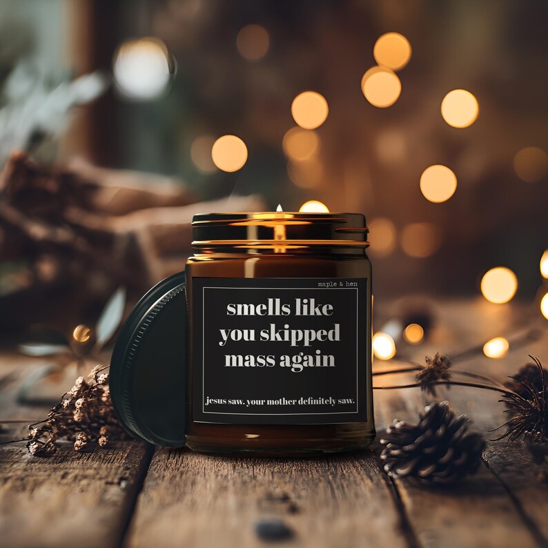 May include: A brown glass jar candle with a black label that reads "smells like you skipped mass again." The candle has a gold lid and is sitting on a wooden surface. The text "jesus saw, your mother definitely saw" is below the main text.