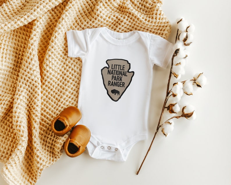 May include: A white short-sleeved baby bodysuit featuring a tan arrowhead-shaped graphic with the black text "LITTLE NATIONAL PARK RANGER" and a small black bison silhouette. The garment has a round neckline and three metal snaps at the bottom.