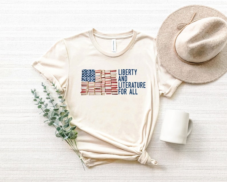 Puede incluir: Camiseta color crema con un dise&ntilde;o de la bandera estadounidense hecha de libros. El texto "LIBERTY AND LITERATURE FOR ALL" est&aacute; impreso en azul marino. Un sombrero beige y una taza blanca est&aacute;n junto a la camiseta.