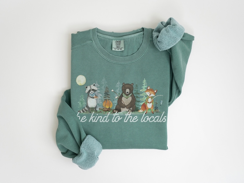 May include: A sage green crewneck sweatshirt featuring a graphic print of a raccoon, a bear, and a fox in a forest setting with a campfire and the text "be kind to the locals" written in white script lettering across the bottom of the illustration.