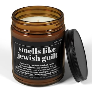 May include: A brown glass jar candle with a black label featuring the white text 'smells like jewish guilt' and additional descriptive copy. The candle has a white wax fill, a single cotton wick, and a separate black metal lid resting beside it.