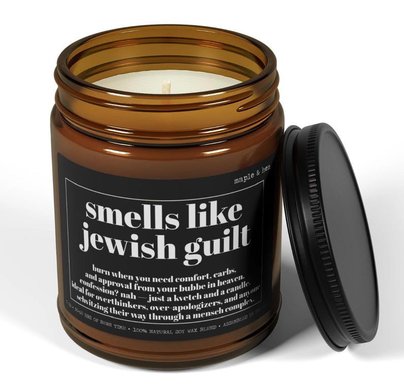 May include: A brown glass jar candle with a black label featuring the white text 'smells like jewish guilt' and additional descriptive copy. The candle has a white wax fill, a single cotton wick, and a separate black metal lid resting beside it.