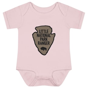 May include: A light pink short-sleeved bodysuit featuring a tan arrowhead-shaped graphic on the chest. The graphic contains the black text "LITTLE NATIONAL PARK RANGER" and a small black silhouette of a bison centred below the lettering.