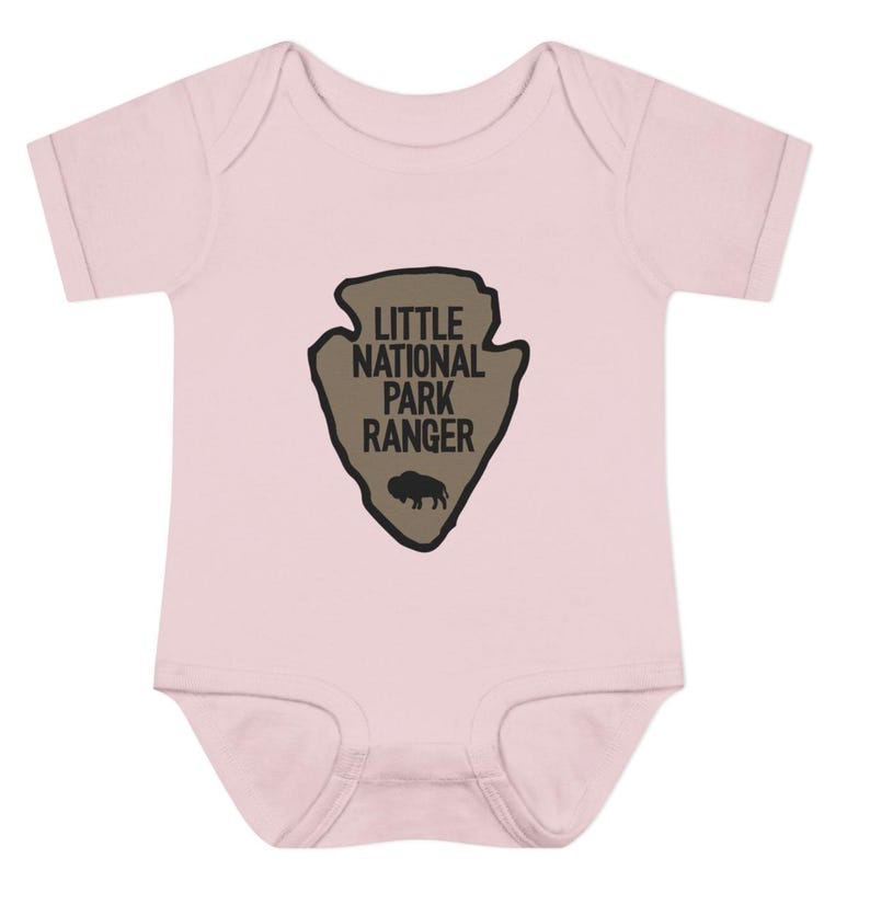 May include: A light pink short-sleeved bodysuit featuring a tan arrowhead-shaped graphic on the chest. The graphic contains the black text "LITTLE NATIONAL PARK RANGER" and a small black silhouette of a bison centred below the lettering.