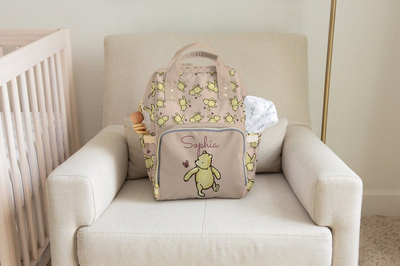 May include: A beige diaper bag with a Winnie the Pooh design, featuring a repeating pattern of yellow Pooh bears. The bag has a front pocket with the name "Sophia" and a Pooh bear graphic. A wooden toy and a white cloth are in the side pockets.