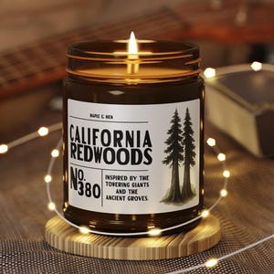 May include: A lit candle in a brown glass jar with a label that reads "CALIFORNIA REDWOODS." The label features two illustrated trees and the text "Inspired by the towering giants and the ancient groves." The candle sits on a wooden coaster, surrounded by string lights.