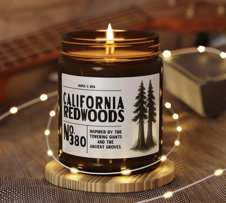 May include: A lit candle in a brown glass jar with a label that reads "CALIFORNIA REDWOODS." The label features two illustrated trees and the text "Inspired by the towering giants and the ancient groves." The candle sits on a wooden coaster, surrounded by string lights.