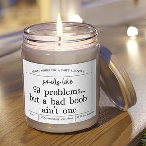 May include: A lit candle in a clear glass jar with a silver lid. The white label features the text: Breast wishes for a swift recovery. Smells like 99 problems... but a bad boob ain't one. It also lists 9 oz, 100% natural soy wax blend, and 50-60 hrs burn time.