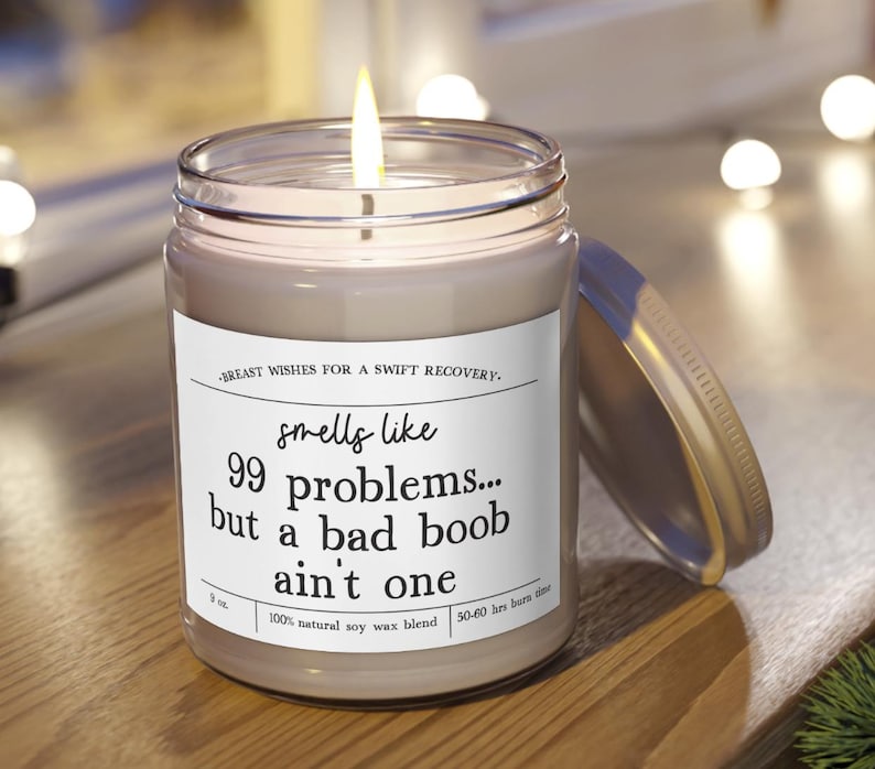 May include: A lit candle in a clear glass jar with a silver lid. The white label features the text: Breast wishes for a swift recovery. Smells like 99 problems... but a bad boob ain't one. It also lists 9 oz, 100% natural soy wax blend, and 50-60 hrs burn time.