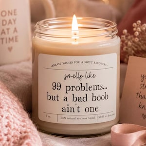 May include: A lit soy wax candle in a clear glass jar with a white label featuring the text: BREAST WISHES FOR A SWIFT RECOVERY. smells like 99 problems... but a bad boob ain't one. The label also states 9 oz. and 50-60 hrs burn time.