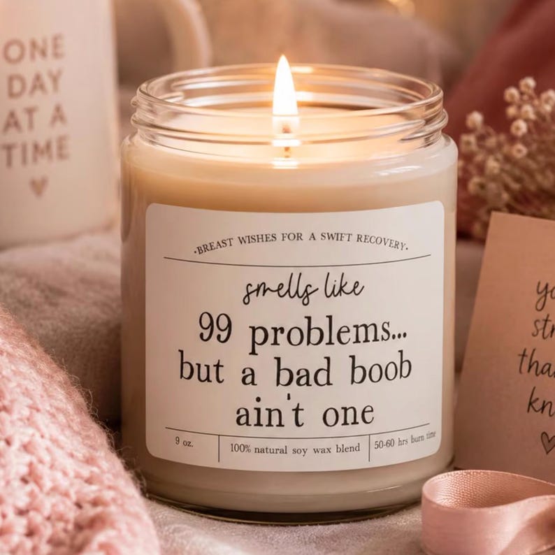 May include: A lit soy wax candle in a clear glass jar with a white label featuring the text: BREAST WISHES FOR A SWIFT RECOVERY. smells like 99 problems... but a bad boob ain't one. The label also states 9 oz. and 50-60 hrs burn time.