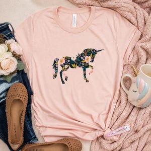 Puede incluir: Una camiseta rosa claro con un dise&ntilde;o de unicornio floral. El unicornio est&aacute; relleno con un patr&oacute;n floral verde oscuro con flores rosas y amarillas. La camiseta est&aacute; combinada con vaqueros, zapatos y una taza de unicornio.