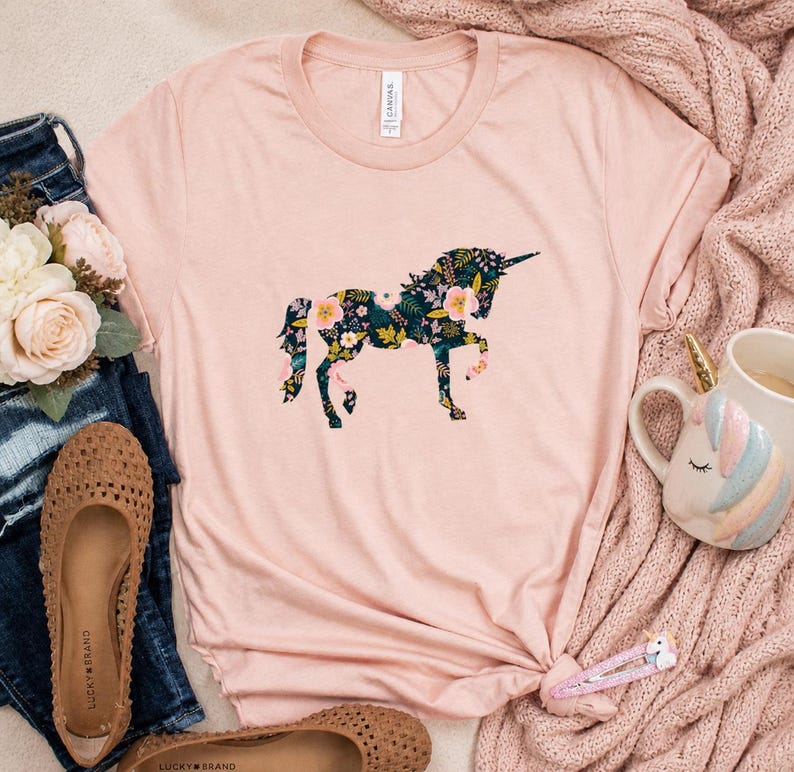Puede incluir: Una camiseta rosa claro con un dise&ntilde;o de unicornio floral. El unicornio est&aacute; relleno con un patr&oacute;n floral verde oscuro con flores rosas y amarillas. La camiseta est&aacute; combinada con vaqueros, zapatos y una taza de unicornio.