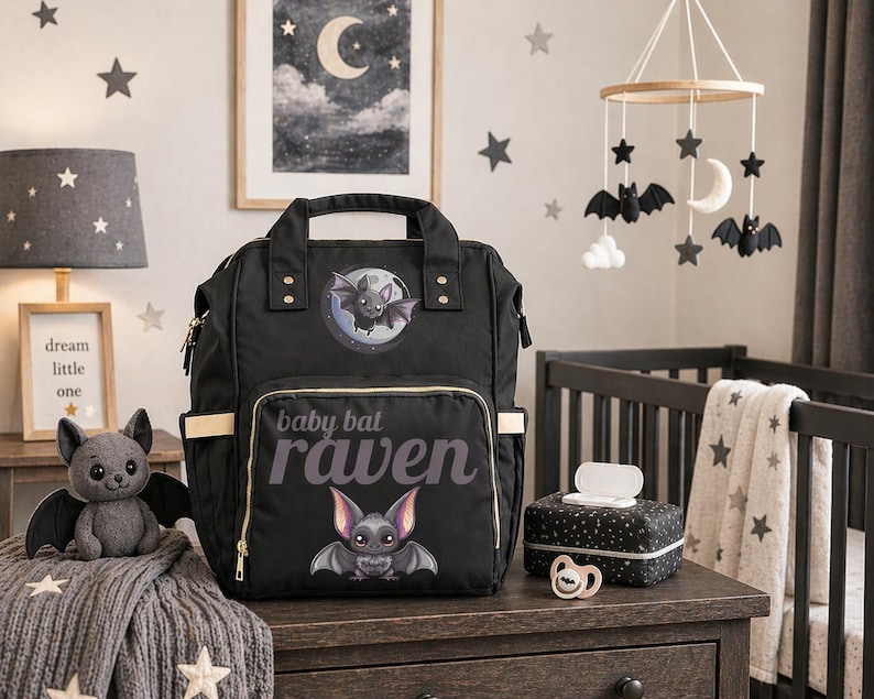 May include: Black baby diaper bag with a bat design and the text "baby bat raven". The bag has multiple pockets and a top handle. A plush bat toy and a matching wipe dispenser are also visible.