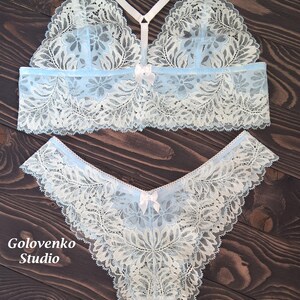 May include: A light blue and white lace bralette and matching panty set featuring floral patterns, thin white straps, and small white bows at the centre of each piece. The text Golovenko Studio is printed in white on the lower left side.