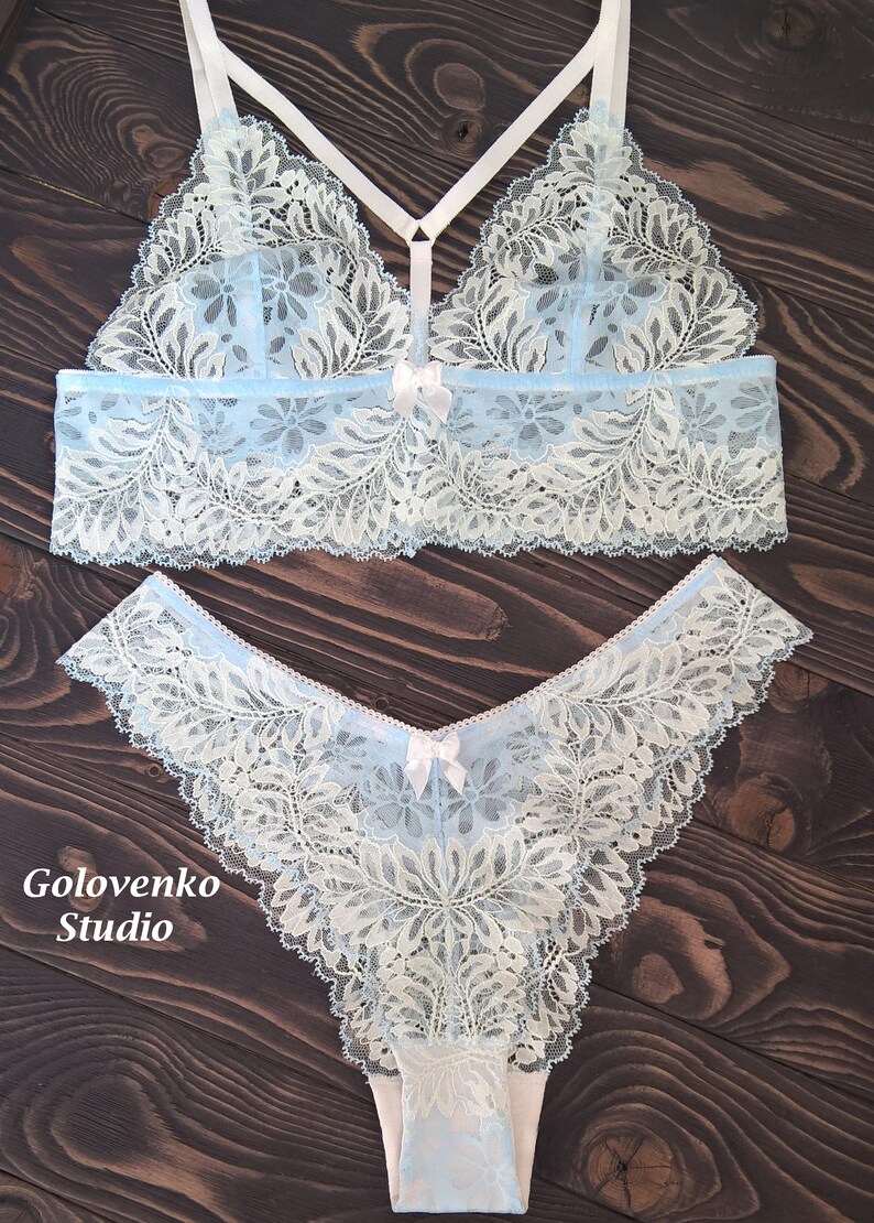 May include: A light blue and white lace bralette and matching panty set featuring floral patterns, thin white straps, and small white bows at the centre of each piece. The text Golovenko Studio is printed in white on the lower left side.