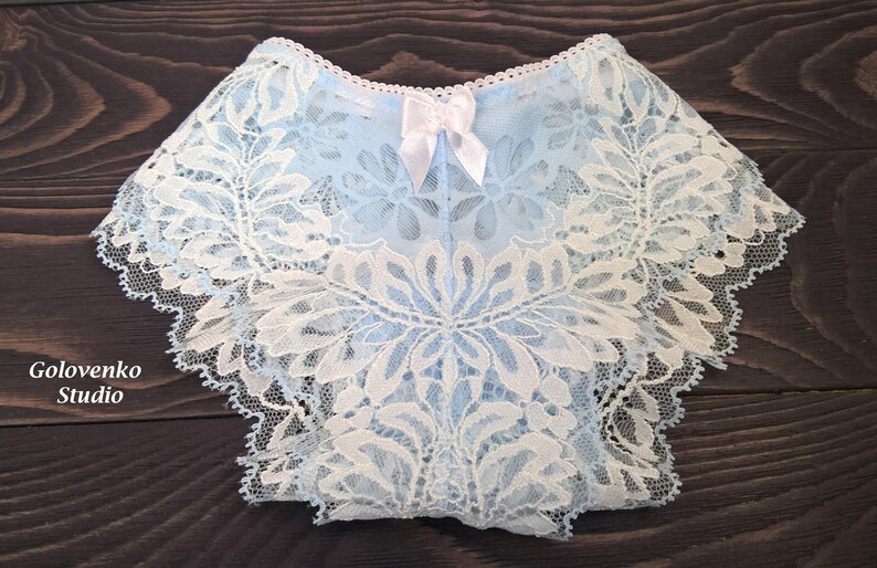 May include: Light blue underwear featuring an intricate white floral lace overlay, a scalloped edge along the waistband and leg openings, and a small white satin bow centred at the top. The text Golovenko Studio is visible on the left side.