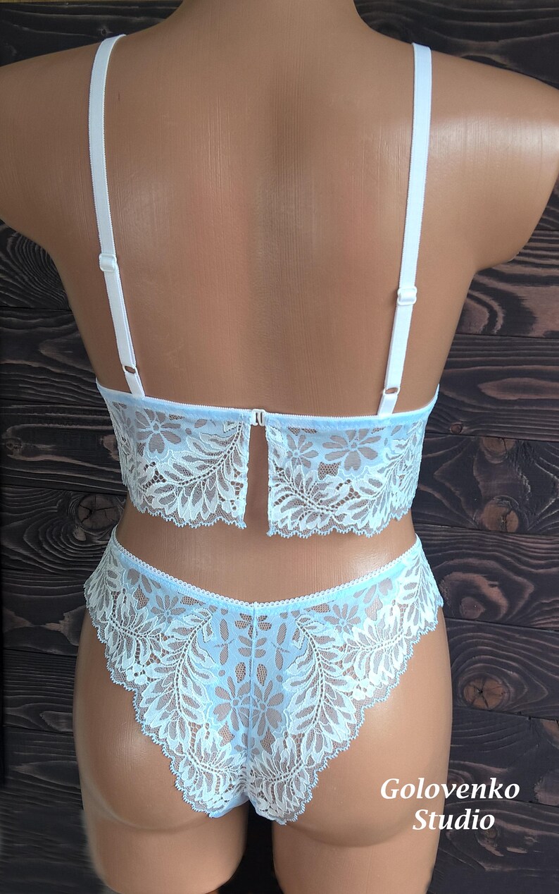 May include: A light blue lace lingerie set consisting of a bralette with adjustable white shoulder straps and matching high-cut knickers. The fabric features a detailed floral pattern with scalloped edges throughout the entire garment set.