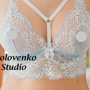 May include: A light blue lace bralette featuring white floral patterns, a central white bow, and white elastic straps that connect at the centre. The text Golovenko Studio is printed on the left side of the garment in a black serif font.