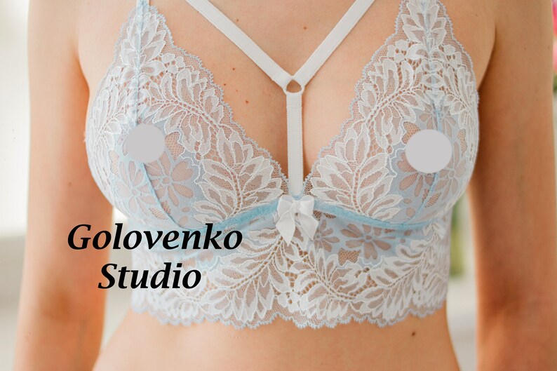 May include: A light blue lace bralette featuring white floral patterns, a central white bow, and white elastic straps that connect at the centre. The text Golovenko Studio is printed on the left side of the garment in a black serif font.