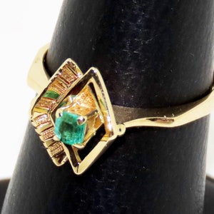 May include: A gold ring featuring a square emerald gemstone set within a geometric design. The ring's band is a simple, polished gold, and the emerald is a vibrant green. The ring is displayed on a black ring holder.