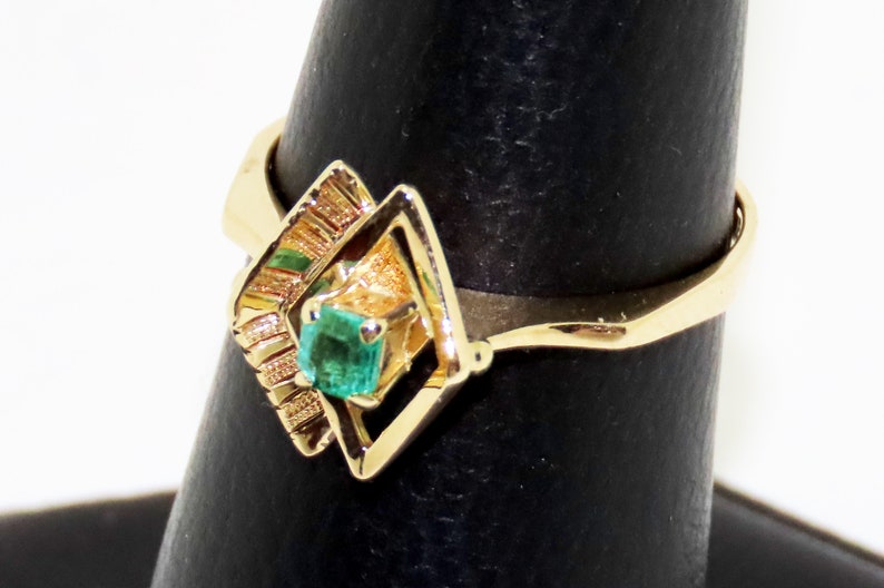 May include: A gold ring featuring a square emerald gemstone set within a geometric design. The ring's band is a simple, polished gold, and the emerald is a vibrant green. The ring is displayed on a black ring holder.