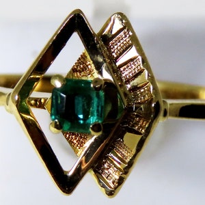 May include: A gold ring featuring a rectangular emerald gemstone set within a geometric design. The ring's setting includes a diamond-shaped frame and textured accents, showcasing a classic and elegant style.