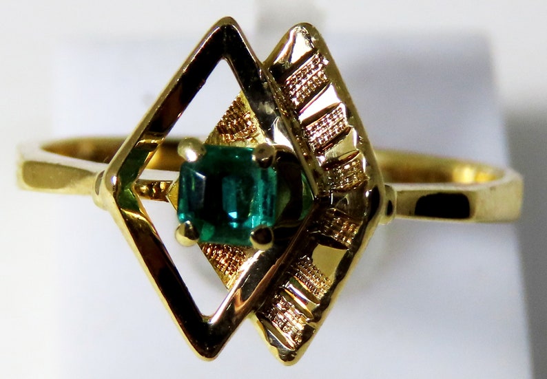 May include: A gold ring featuring a rectangular emerald gemstone set within a geometric design. The ring's setting includes a diamond-shaped frame and textured accents, showcasing a classic and elegant style.