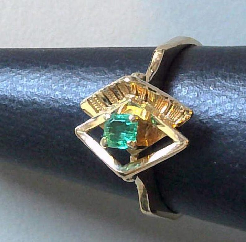 May include: A gold ring featuring a square emerald gemstone set within a geometric design. The ring's setting has a textured, ornate pattern, and the band is a simple, polished gold. The emerald is a vibrant green color.