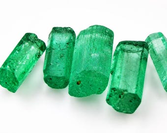 Puede incluir: Cinco cristales de esmeralda en bruto y alargados, con un tono verde vibrante y calidad translúcida. Cada piedra presenta una estructura prismática hexagonal con texturas superficiales naturales, inclusiones internas y longitudes variables.