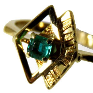 May include: A gold ring featuring a square-cut emerald gemstone set within a geometric, openwork design. The ring's band and setting are a polished gold color, with intricate detailing around the emerald.