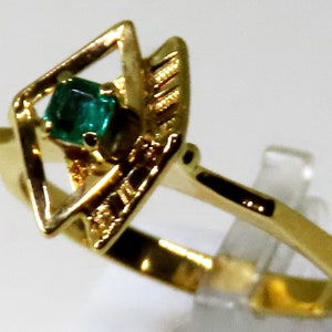 May include: A gold ring featuring a square-cut emerald gemstone set within a geometric design. The ring has a polished gold band and decorative accents around the emerald. The ring is displayed on a clear stand.