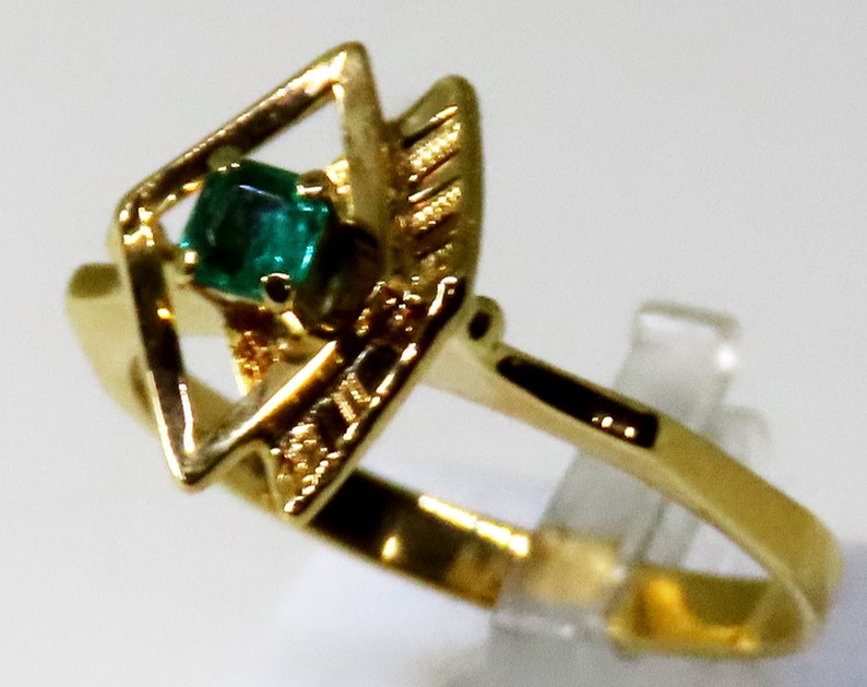 May include: A gold ring featuring a square-cut emerald gemstone set within a geometric design. The ring has a polished gold band and decorative accents around the emerald. The ring is displayed on a clear stand.