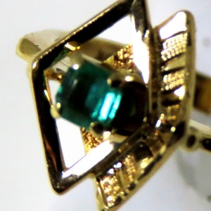 May include: Close-up of a gold ring featuring a square-shaped setting with a vibrant green gemstone. The ring has intricate gold detailing around the gemstone and a classic band. The ring is a piece of jewelry.