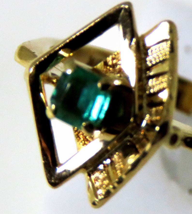 May include: Close-up of a gold ring featuring a square-shaped setting with a vibrant green gemstone. The ring has intricate gold detailing around the gemstone and a classic band. The ring is a piece of jewelry.