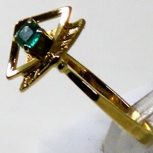 May include: A gold ring featuring a square-cut, emerald-green gemstone set within a geometric design. The ring's band is a simple, polished gold, and the setting has decorative elements.