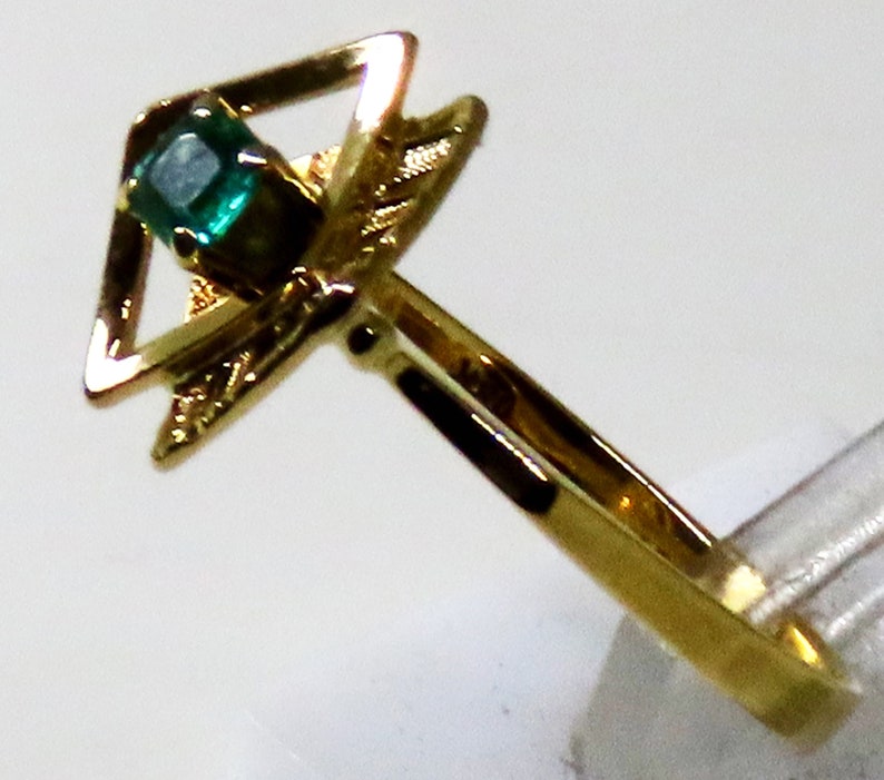 May include: A gold ring featuring a square-cut, emerald-green gemstone set within a geometric design. The ring's band is a simple, polished gold, and the setting has decorative elements.