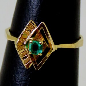 May include: A gold ring featuring a square emerald gemstone set within a geometric design. The ring has a unique, openwork setting with a diamond-like shape and decorative elements on the side.