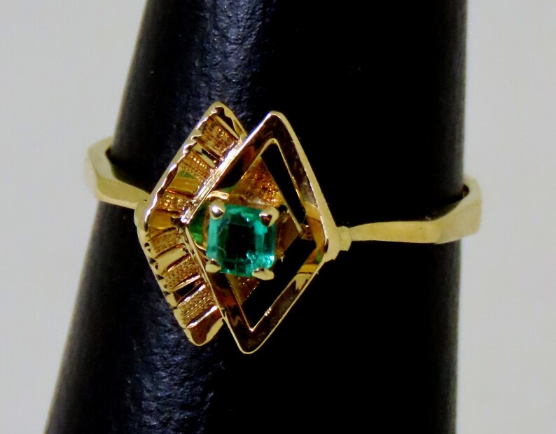 May include: A gold ring featuring a square emerald gemstone set within a geometric design. The ring has a unique, openwork setting with a diamond-like shape and decorative elements on the side.