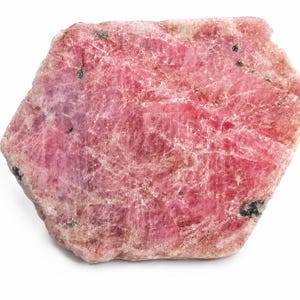 Puede incluir: Un espécimen mineral en bruto de forma hexagonal que presenta una superficie cristalina de color rosa y magenta vibrante con vetas blancas visibles y pequeñas inclusiones de color gris oscuro dispersas por la cara texturizada e irregular.