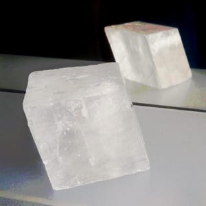 Może przedstawiać: P&oacute;łprzezroczysty, biały, sześcienny okaz minerału o krystalicznej, teksturowanej powierzchni. Obiekt znajduje się na pierwszym planie, podczas gdy drugi, nieco rozmyty, podobny sześcienny kawałek minerału jest widoczny w tle.