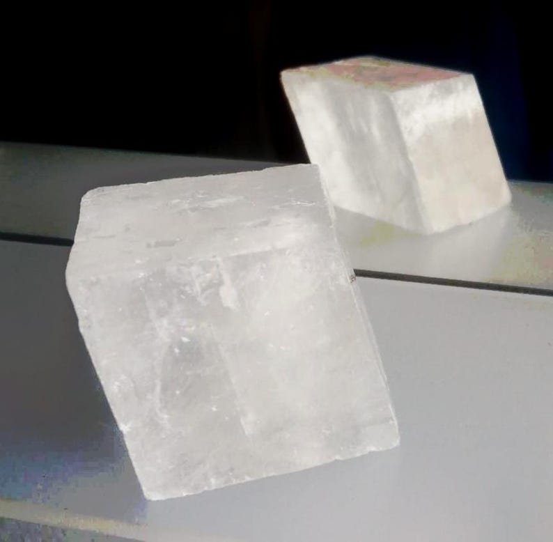 Może przedstawiać: P&oacute;łprzezroczysty, biały, sześcienny okaz minerału o krystalicznej, teksturowanej powierzchni. Obiekt znajduje się na pierwszym planie, podczas gdy drugi, nieco rozmyty, podobny sześcienny kawałek minerału jest widoczny w tle.