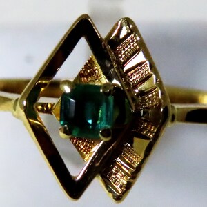 May include: A gold ring featuring a rectangular, emerald-green gemstone set within a decorative, openwork design. The ring's band and setting are a polished gold color, with a geometric pattern on the sides of the gemstone.