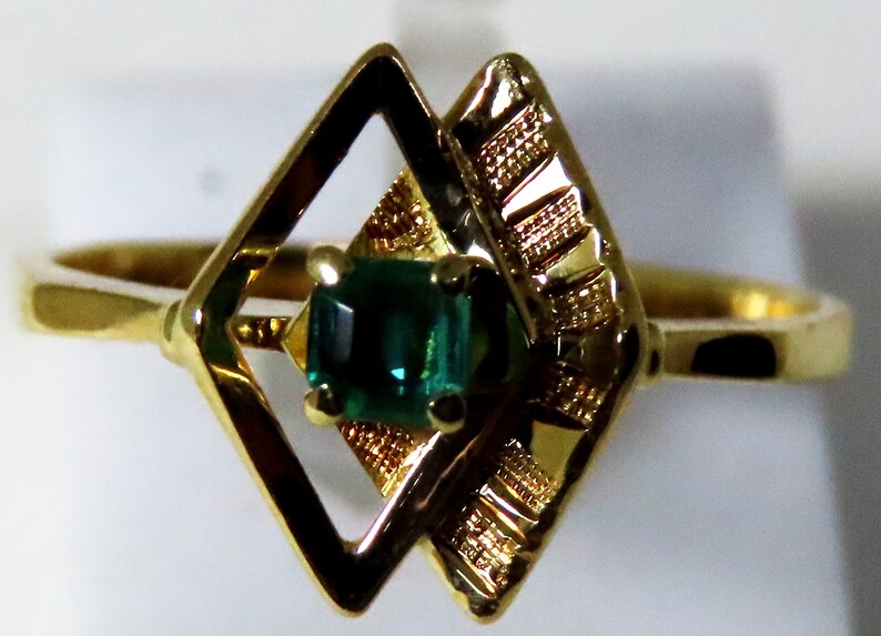 May include: A gold ring featuring a rectangular, emerald-green gemstone set within a decorative, openwork design. The ring's band and setting are a polished gold color, with a geometric pattern on the sides of the gemstone.