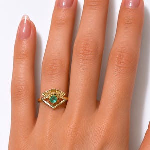 May include: A gold ring featuring a square emerald gemstone set within a geometric design. The ring has a fan-like detail above the emerald and is worn on a finger. The ring is a piece of jewelry.