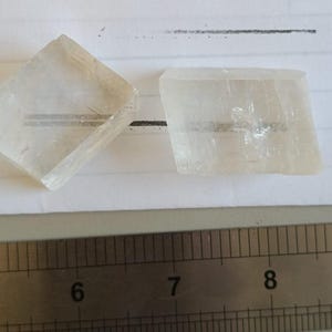 Może przedstawiać: Dwa przezroczyste, romboidalne kryształy kalcytu z widocznymi wewnętrznymi pęknięciami i gładkimi powierzchniami. Metalowa linijka umieszczona pod kryształami wskazuje, że lewy kryształ ma 2 cm, a prawy kryształ 2,5 cm szerokości.