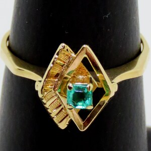 May include: A gold ring featuring a square emerald gemstone set within a diamond-shaped frame. The ring has a textured design on one side and a smooth, polished band. The emerald is a vibrant green color.